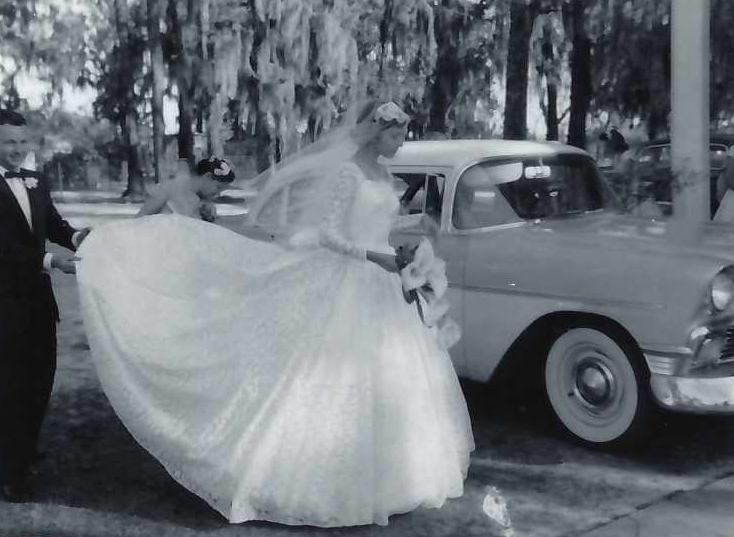 A black and white photo of a bride and groom standing together in front of a vintage car, smiling joyfully